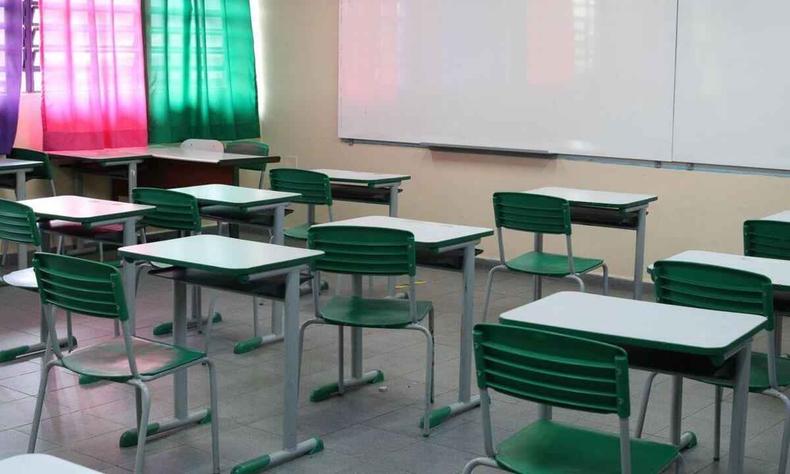 Vista de sala de aula vazia. Foto tirada do fundo da sala. No fundo da imagem  possvel ver um quadro branco e,  esquerda, janelas que esto tampadas com cortinas coloridas. 