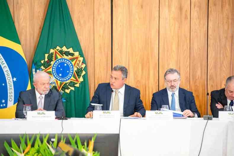 Lula e Rui Costa sentados a mesa coberta com toalha de mesa branca em evento oficial com a bandeira do Brasil ao fundo 