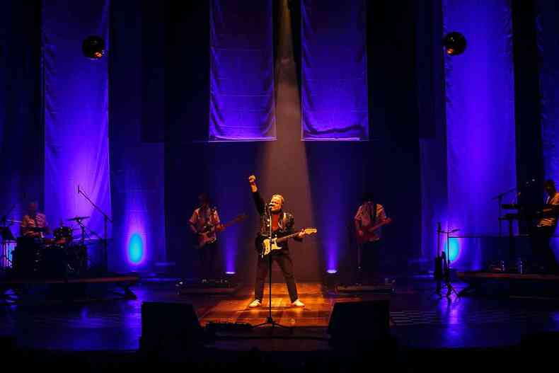 Leo Jaime, de roupa preta e sapato branco, segura baixo com a mo esquerda e ergue a direita no palco, em frente a microfone