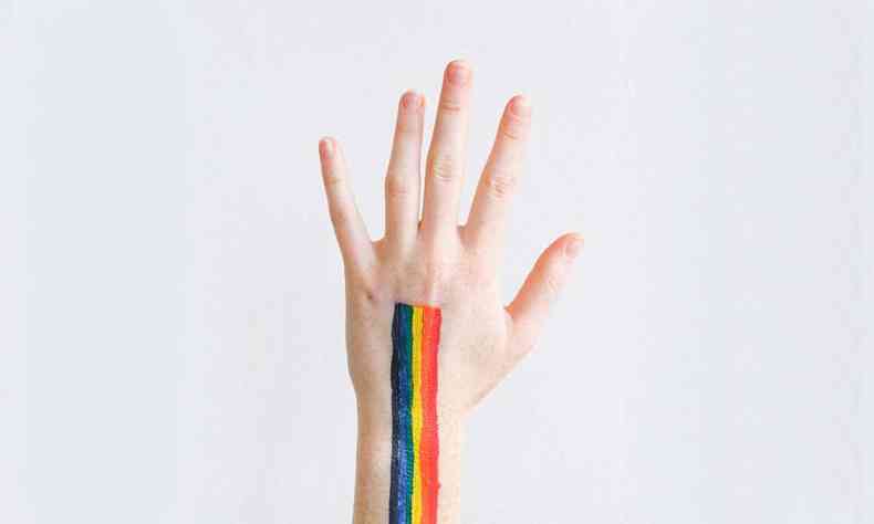 Mão de uma pessoa branca levantada, com as cores da bandeira LGBT pintadas no dorso