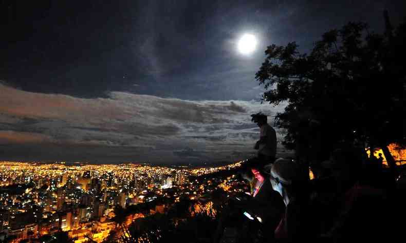Cidade iluminada por eletricidade, lua no céu e jovens sentados observando