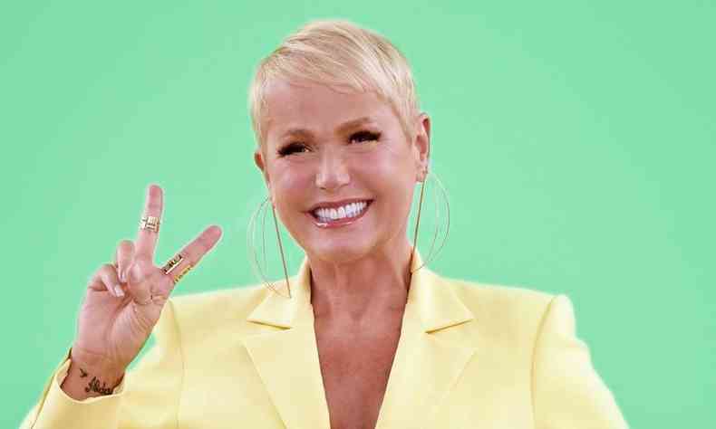 Xuxa com uma roupa amarela fazendo o sinal de dois com os dedos, em um fundo verde