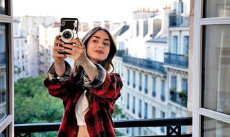 Lily Collins vive a jovem americana enfeitiçada pela França(foto: Stephanie Branchu/Netflix) Lily Collins faz selfie tendo ao fundo paisagem de Paris em cena da série Emily em Paris