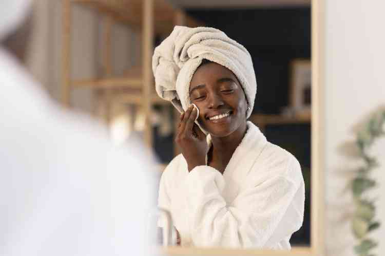 Mulher negra com talhas brancas no corpo e na cabeça de frente para o espelho sorrindo fazendo seu cuidado com a pele