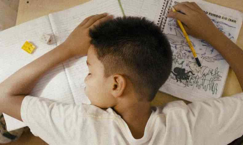 Imagem é um take do filme 'Picolé'. Nela, há um menino negro deitado sobre cadernos e atividades escolares. Ele usa uma camiseta branca e segura um lápis amarelo