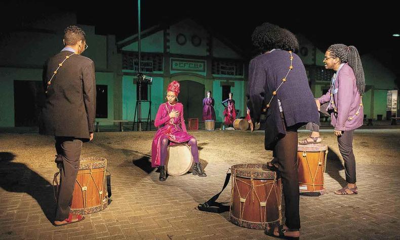 Atriz est sentada em tambor e atores seguram tambores no espetculo teatral Alm do rio