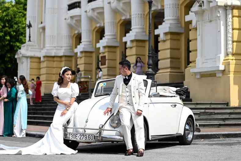 Vestidos de branco e apoiados num Fusca, noiva e noivo se olham em ensaio de casamento