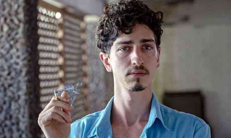 de camisa azul, o ator Johnny Massaro ergue um cigarro com a mo direita 