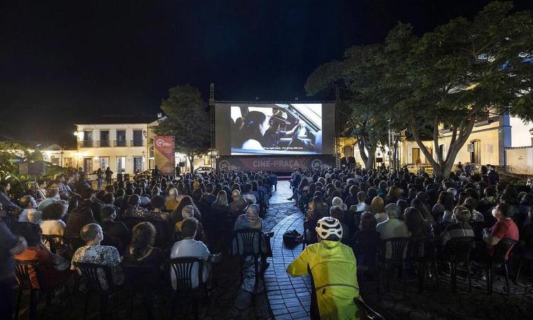 Pblico sentado em cadeiras assiste a filme em tela montada ao ar livre na cidade histrica de Tiradentes