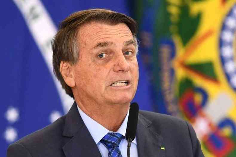 Bolsonaro com cara de irritado com Bandeira do Brasil ao fundo 