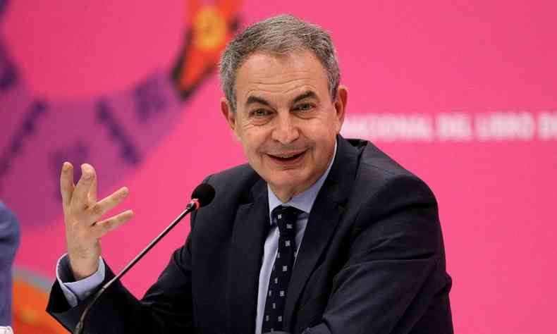 Jos Luis Rodrguez Zapatero usando terno preto sentado a mesa com um microfone a sua frente em um fundo rosa 