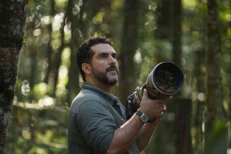 De camisa verde, de p, em ambiente florestal, o fotgrafo Joo Marcos Rosa empunha cmera com lente de grande alcance