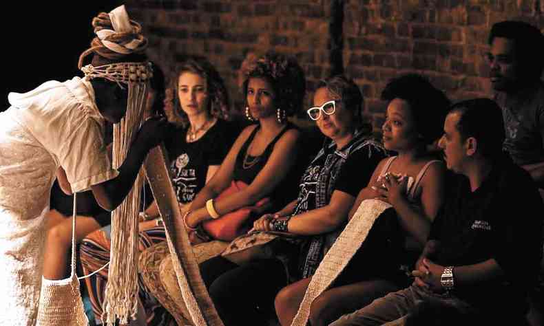 Pessoas sentadas em cadeiras observam atriz vestida de branco e com turbante em cena do espetculo  sombra da goiabeira