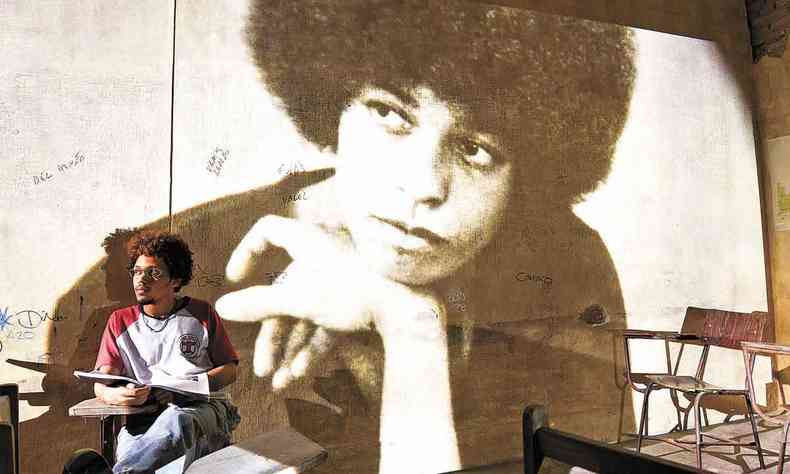 o ator Lucas Limeira sentado em sala de aula com foto da ativista Angela Davis ao fundo, em cena do filme Cabea de Ngo