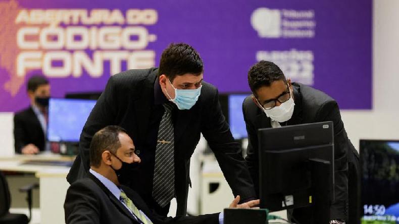 Dois homens brancos e um pardo usam mscaras e olham para a tela de um computador; ao fundo um grande painel est escrito 'abertura do cdigo-fonte'