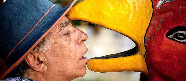 De chapu, o cineasta mineiro Neville D'Almeida posa com o rosto dentro do bico amarelo da escultura de uma ave