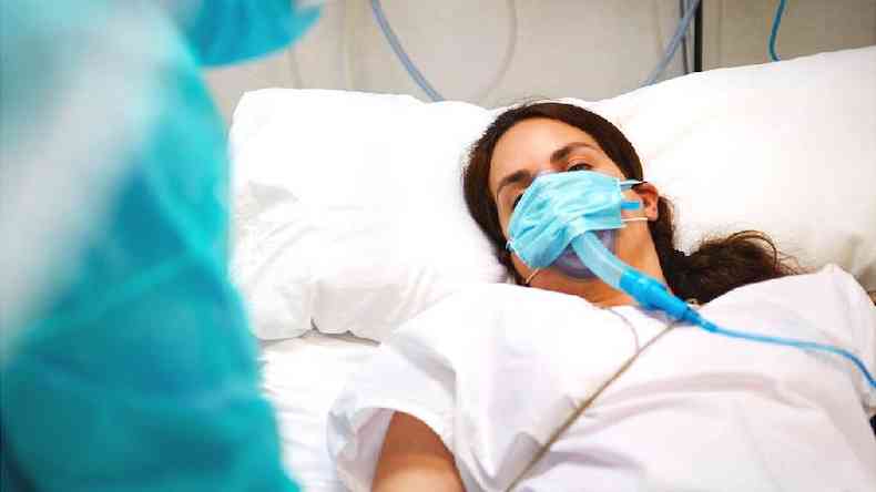 Fotografia colorida mostra mulher branca de cabelo castanho, de máscara, em cama de hospital