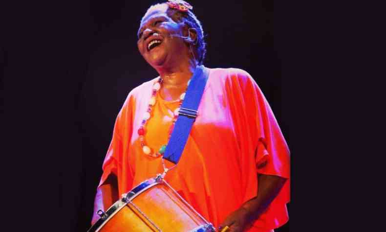 Dona Valdete Cordeiro usando um vestido largo laranja forte tocando tambor