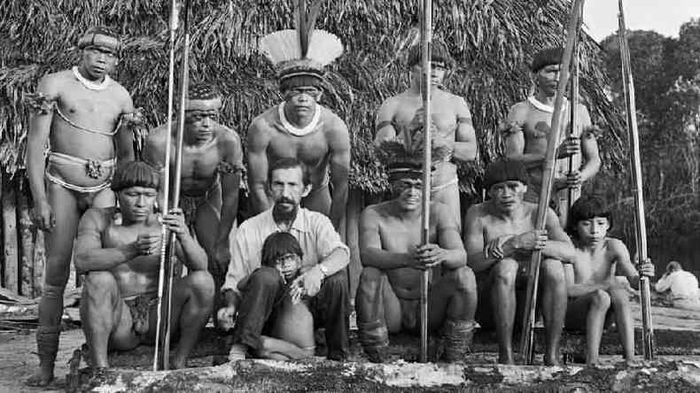 Fotografia em preto e branco mostra 9 homens do povo Yawalapiti, um homem branco e uma criana; parte do grupo est sentada
