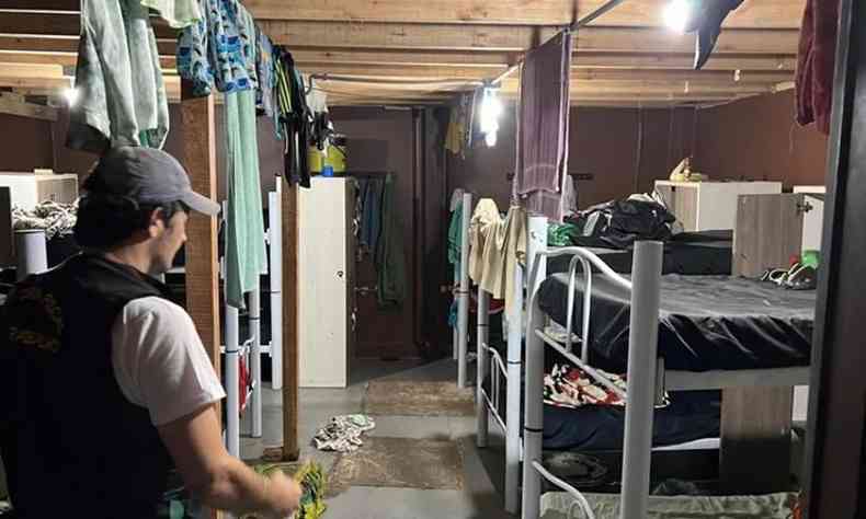 Foto do quarto em que os trabalhadores moravam. H diversas beliches e roupas penduradas em um varal improvisado em frente s camas. Um agente branco da PF usando um bon circula pelo local