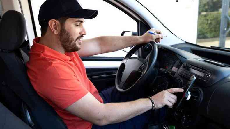 homem sentado em banco do motorista, com camiseta vermelha, mexe em celular posicionado no painel do carro