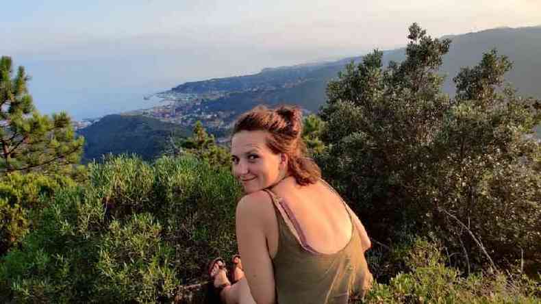 Megan Morgan sentada em uma montanha, olhando por cima do ombro para a cmera. Ao longe,  possvel ver arbustos e o mar