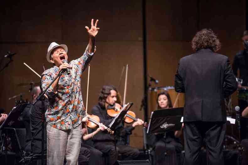Usando chapu, segurando o microfone e com o rosto emocionado, Antonio Nobrega se apresenta ao lado do maestro Rodrigo Toffolo, de costas, que est regendo a Orquestra Ouro Preto