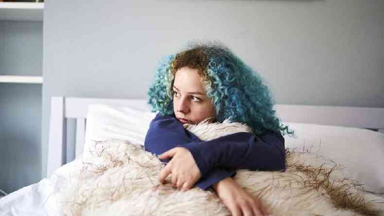 Mulher branca de cabelos encaracolados e tingidos de azul olha para o lado na cama