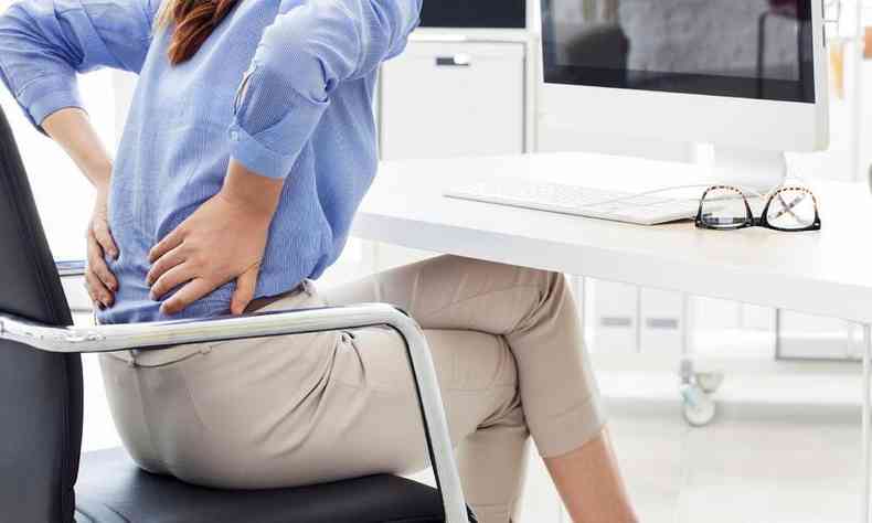 mulher sentada no escritrio com as mos na regio da lombar, indicando dor 