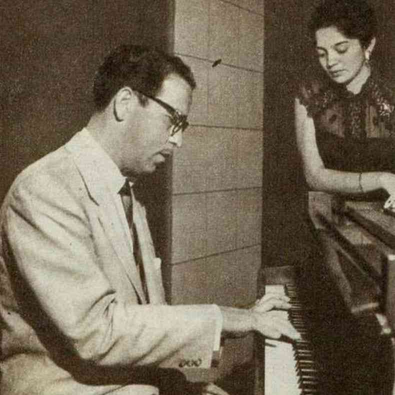 Foto em preto e branco mostra Vadico tocando piano, com mulher ao fundo observando