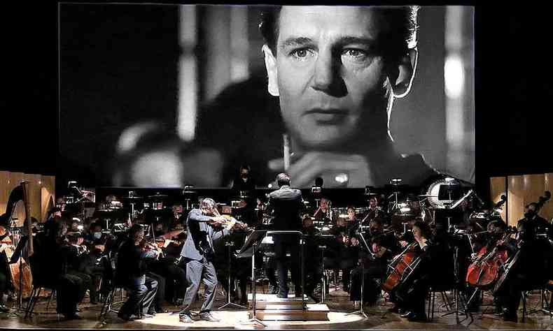 Orquestra Sinfônica de Minas Gerais toca em palco, com imagem de filme projetada ao fundo, em telão