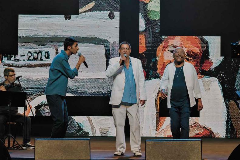 Mosquito, Zeca Pagodinho e Martinho da Vila cantam juntos ''Conselho'', de Adilson Bispo e Zé Roberto, no especial que apresenta um total de 22 músicas (foto: HBO Max / Dueto Filmes / Divulgação) Mosquito, Zeca Pagodinho e Martinho da Vila, em pé no palco, segurando microfones, com cenografia de pinturas modernistas atrás de si