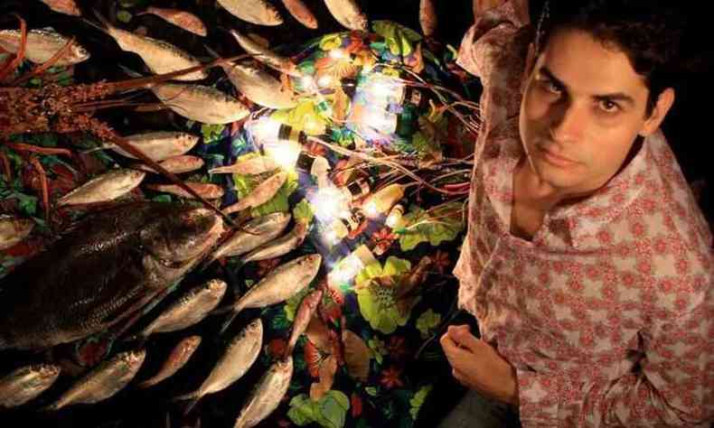 Thiago Amud olha para a cmera, de baixo para cima, tendo a seu lado arranjo iluminado com pequenos peixes mortos