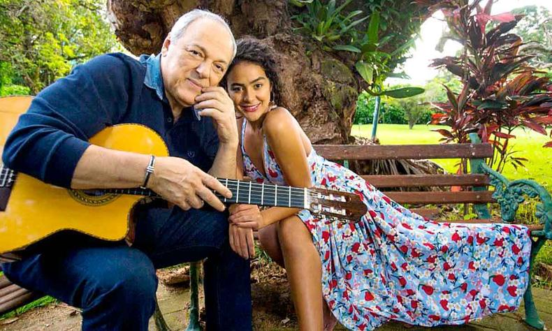 Sentados num banco de jardim, Toquinho segura o violo ao lado da cantora Camilla Faustino, recostada nele, que usa vestido florido 