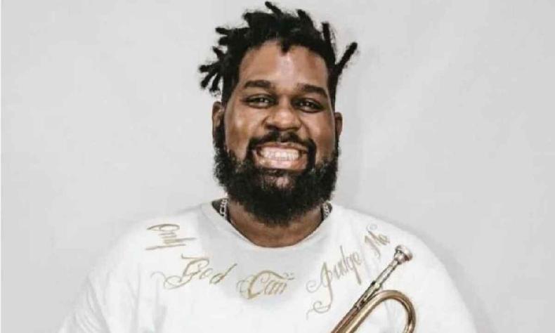 Jeronimo, homem negro, com barba e cabelos com dreads curtos, usa uma blusa branca e sorri para a foto