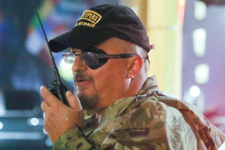 Stewart Rhodes, um homem branco de meia idade, de bigode e bon, usando um tapa olho e um walkie talkie