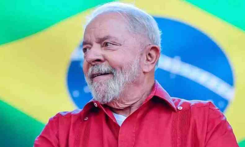 Lula de camisa vermelha sorrindo com uma bandeira do Brasil ao fundo