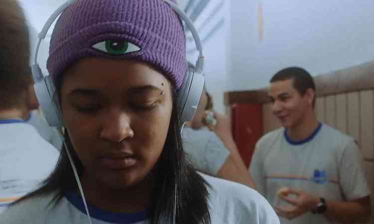 Clara Lima, com fones de ouvido e usando touca roxa, olha para baixo em cena do curta 'Nada', dirigido por Gabriel Martins. Ao fundo v-se um estudante