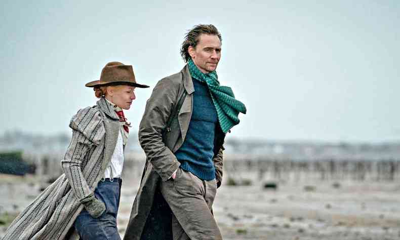 Vestidos com roupas de inverno, os atores Claire Danes e Tom Hiddleston caminham na areia da praia em cena de A serpente de Essex