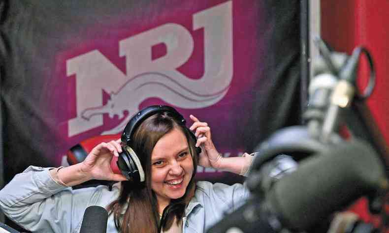 A DJ Yana Manuilova leva as mos aos fones de ouvido que usa em cabine de estdio que tem escrita na parede de fundo, em letras brancas sob fundo vermelho, NRJ
