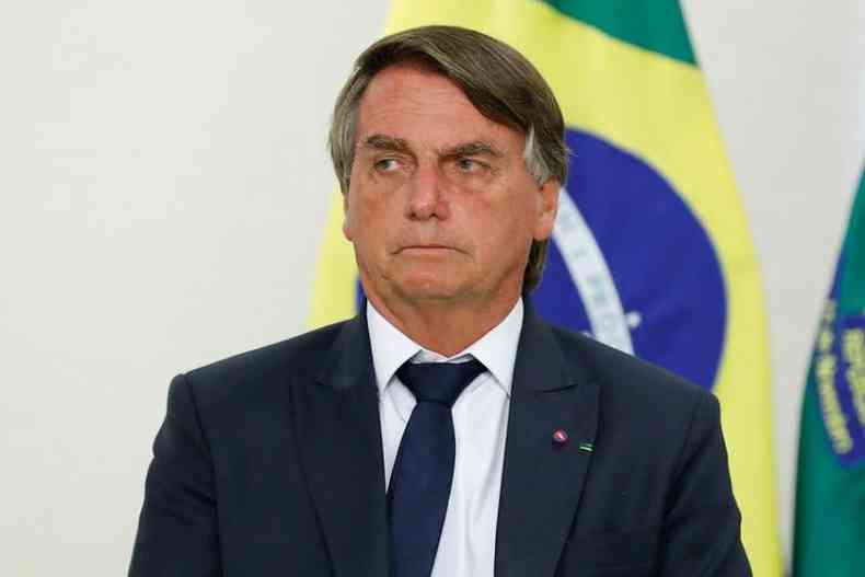 Jair Bolsonaro olha desconfiado para o lado esquerdo. Ele veste um terno preto e uma gravata azul marinho. Atrs dele, uma bandeira do Brasil