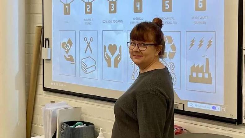 A professora Marika Kerola, uma mulher branca de cabelos castanhos com franja, preso em um coque, e culos; ela est em frente a uma exibio de slides
