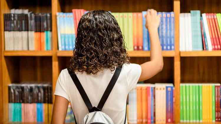 Adolescente de costas e com mochila olhando para livros em prateleiras