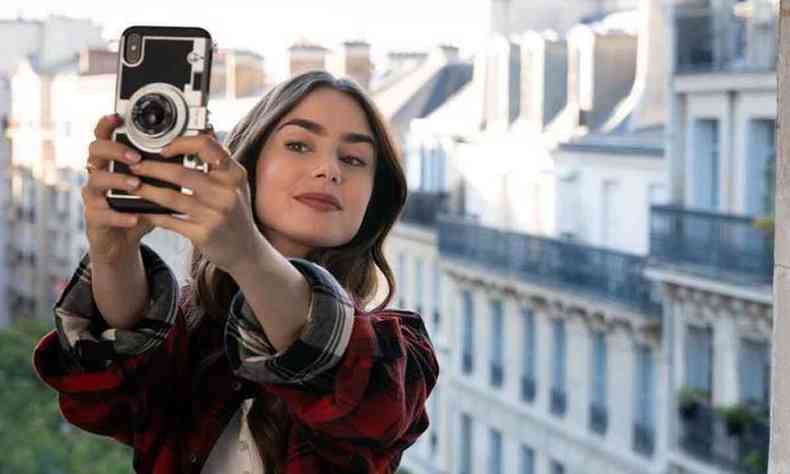 Emily, vivida por Lily Collins, tira selfie, tendo ao fundo casario de Paris 