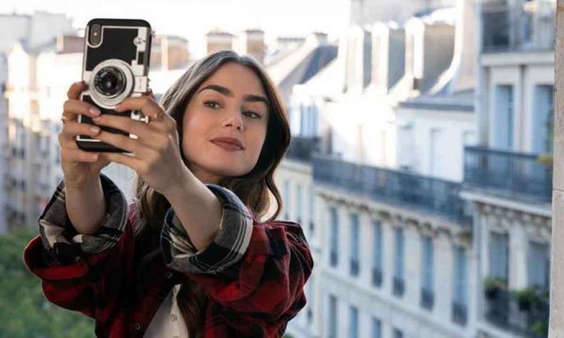 Emily, vivida por Lily Collins, tira selfie, tendo ao fundo casario de Paris 