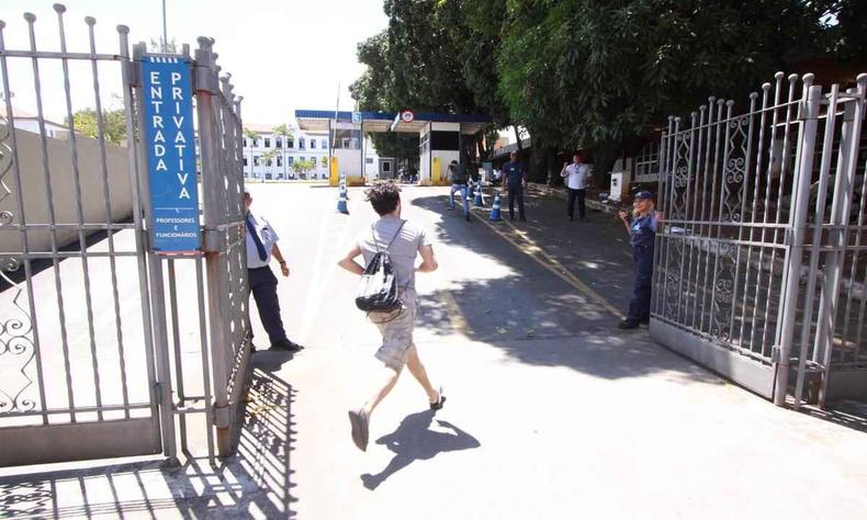 Estudante de blusa cinza, bermuda branca e tnis, correndo para entrar no porto de grade antes que ele se feche