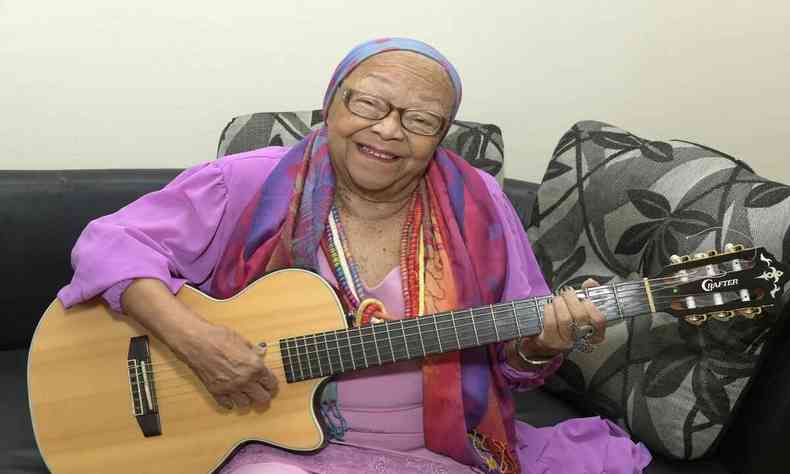 Veterana cantora Dona Jandira posa com seu violo em punho sentada em um sof