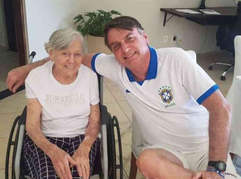 O presidente Jair Bolsonaro ao lado da mãe, Olinda Bolsonaro. Ela está sentada em uma cadeira de rodas.