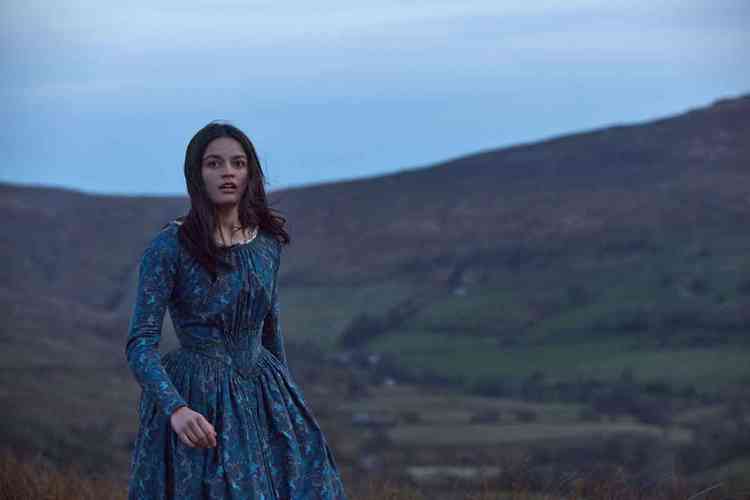A atriz Emma Mackey, de vestido, em paisagem montanhosa, em cena de 'Emily'
