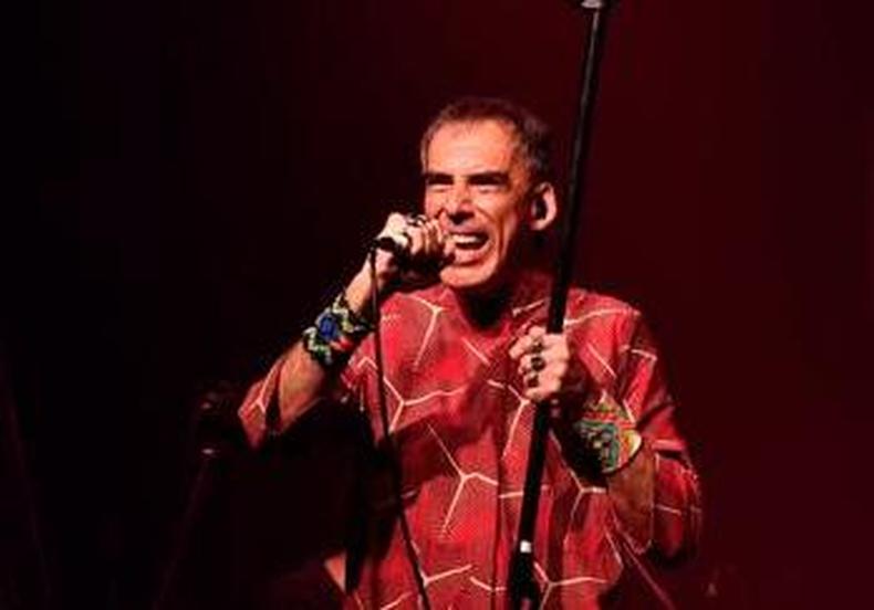 Arnaldo Antunes de vestido estampado, cala cqui e braceletes indgenas, Arnaldo Antunes segura o microfone e sorri